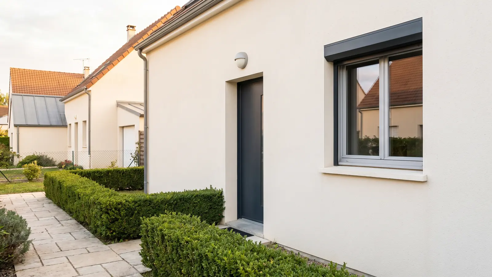 Entrée d’une maison rénovée, porte anthracite, façade enduite et haie taillée (image d’illustration)